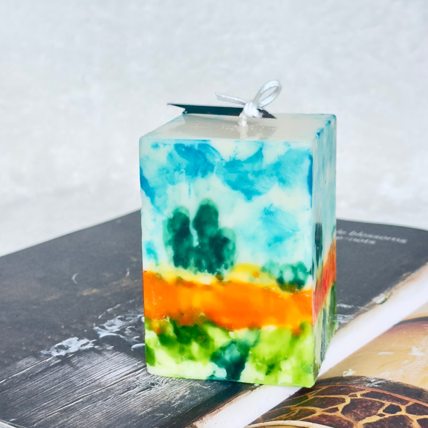 Hand-painted square pillar candle with azure sky, meadow greens, and a saffron-orange horizon band, inspired by Monet’s dawn landscapes.