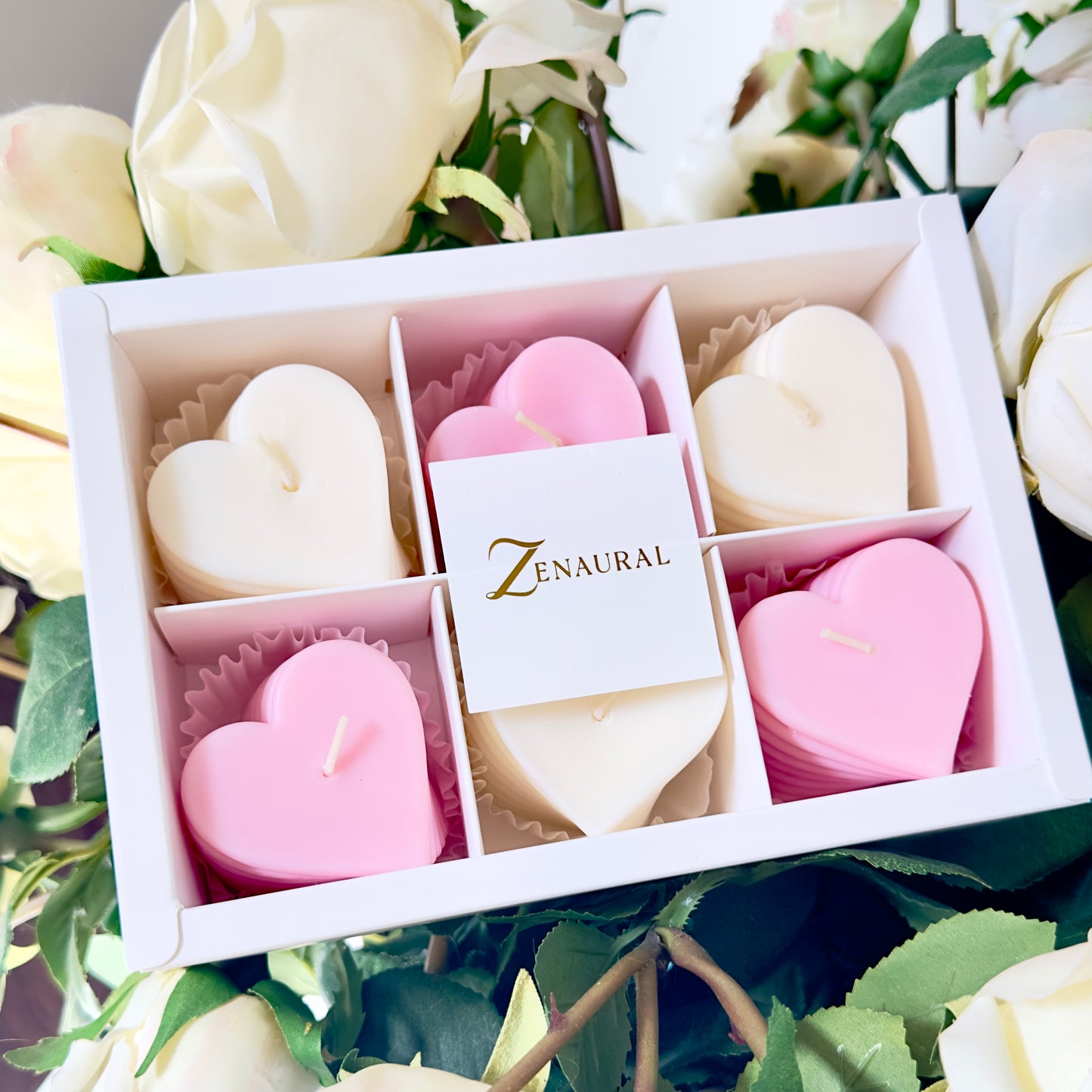 Heart-shaped spiral candles in blush pink and white, arranged in a Zenaural gift box among white roses.