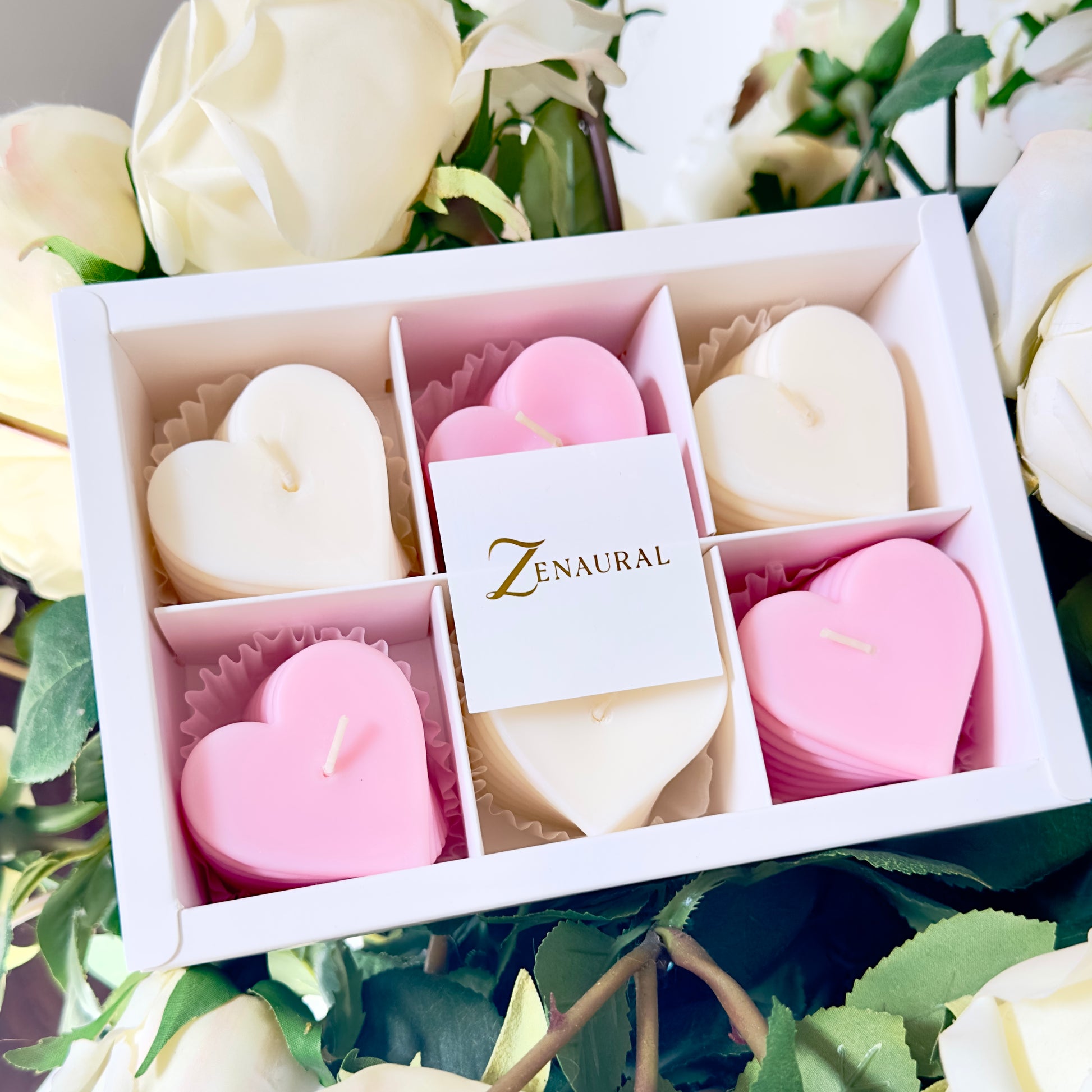Heart-shaped spiral candles in blush pink and white, arranged in a Zenaural gift box among white roses.