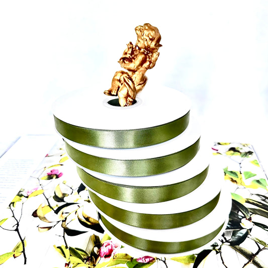 Stacked olive-green satin ribbon spools with a gold cherub on top, set on floral book pages against a white background.
